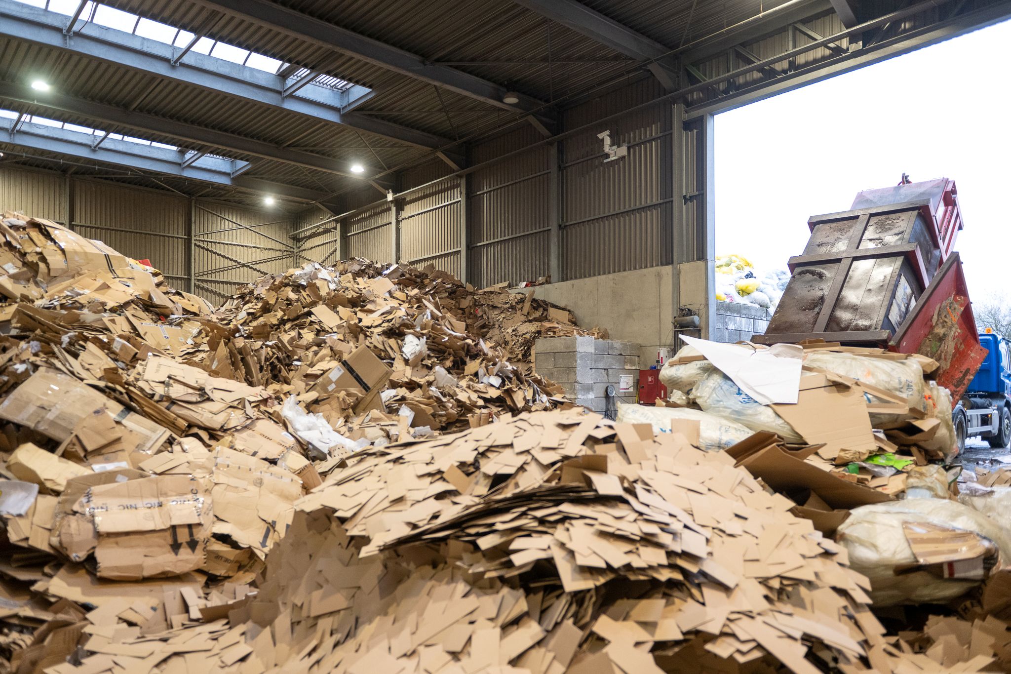 Sfeerbeeld van een afvalverwerkingshal met stapels karton en papier, met een bewakingscamera op de achtergrond
