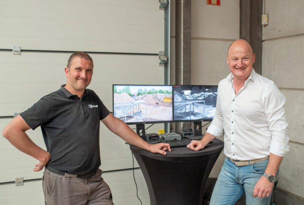 Twee mannen naast twee schermen met live optische en thermische camerabeelden van een site met houtafval