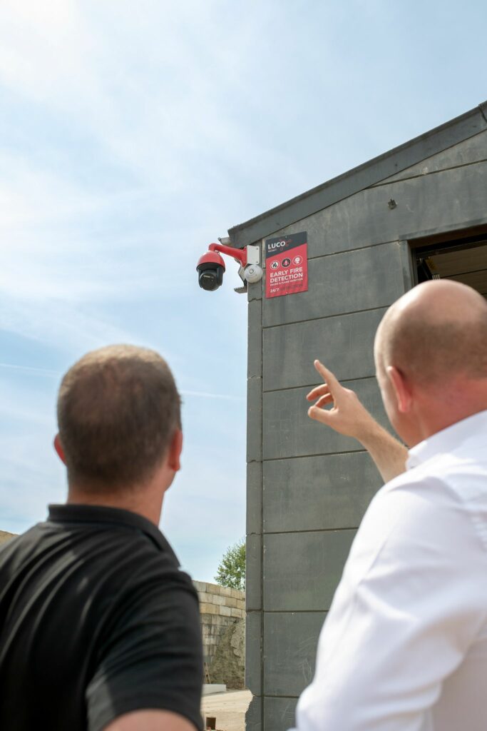 Twee mannen bekijken een LUCO-bewakingscamera met een Early Fire Detection-bordje, gemonteerd op de hoek van een bedrijfsgebouw