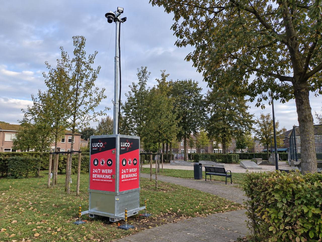 Mobiele LUCO-camerabewakingsunit met camera op een hoge paal en een rood-grijs bord met de tekst '24/7 werfbewaking', opgesteld op een grasveld met bomen, een bankje en woonhuizen op de achtergrond