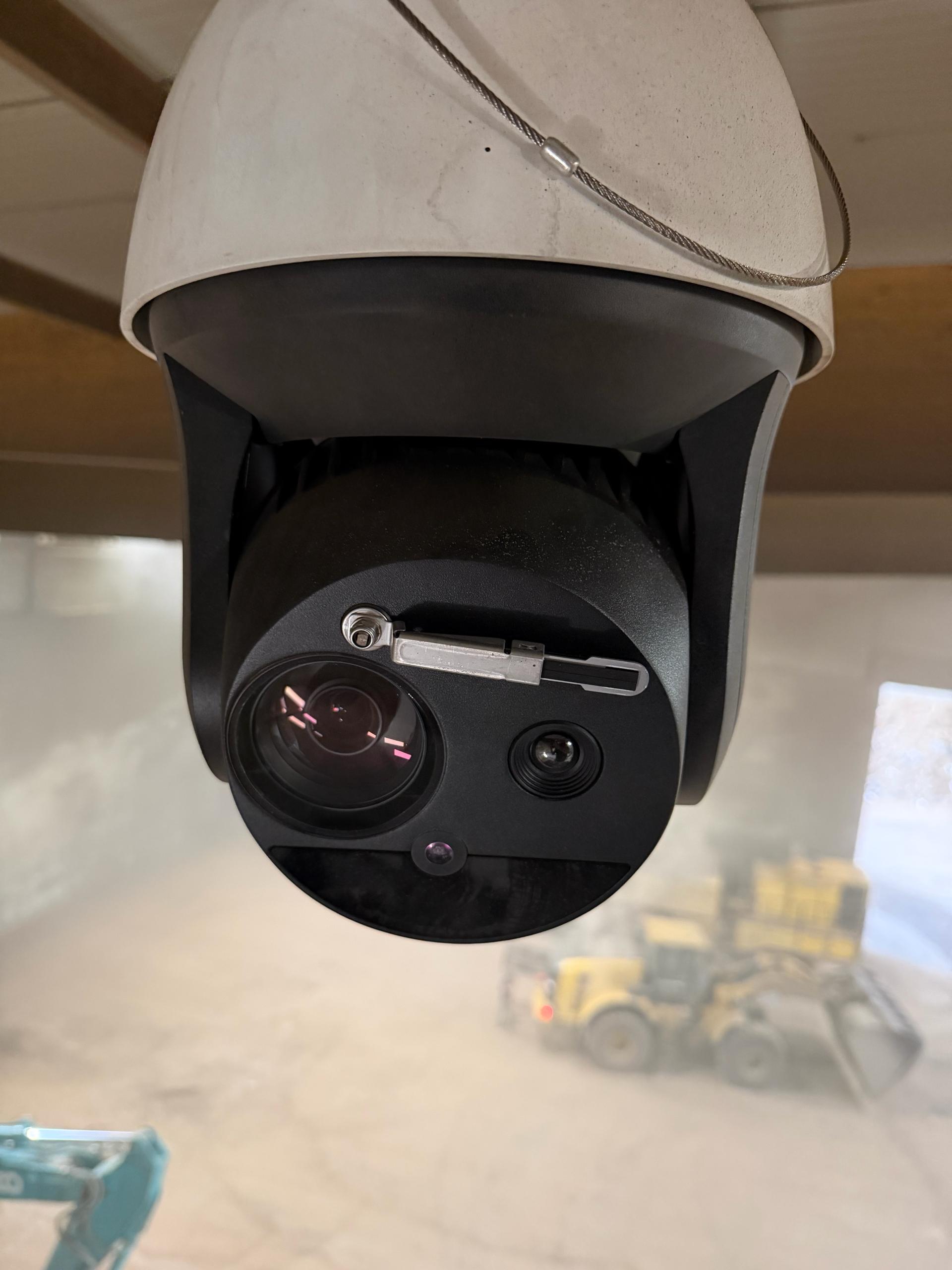 Wit-met-zwarte PTZ-bewakingscamera met dubbele optische lens, plafondgemonteerd aan een metalen kabel boven een magazijn- of werkplaatsruimte